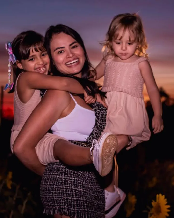 Juliana Martins com as duas filhas no colo, num campo de girassóis ao pôr do sol